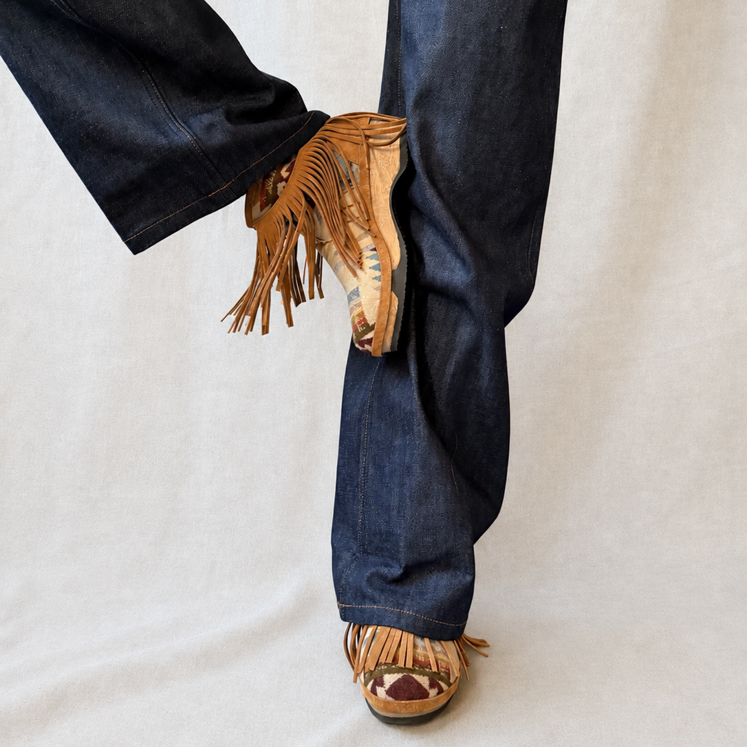 A person wearing dark blue wide-leg jeans and tan fringed clog boots, shown from the waist down against a light neutral fabric backdrop.