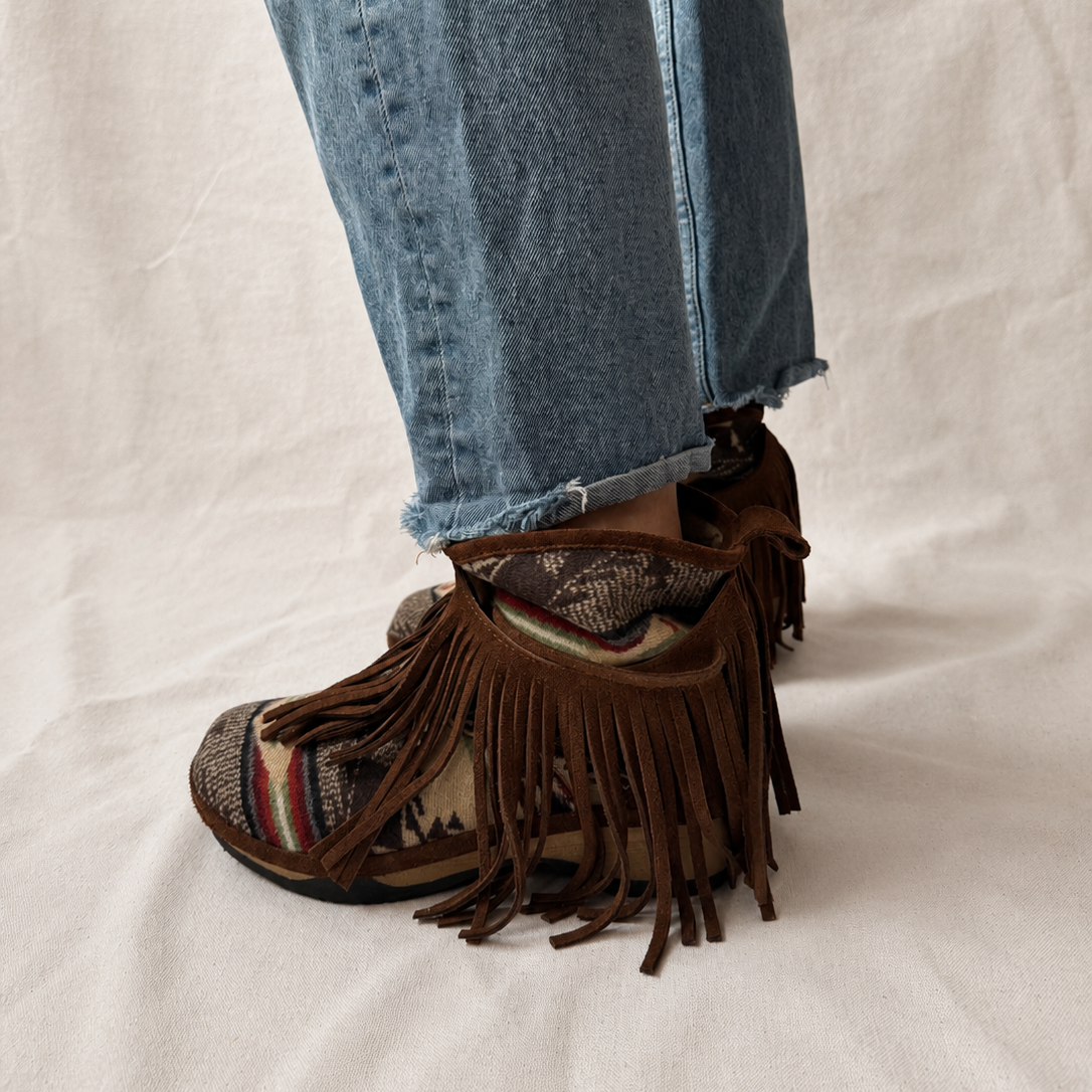 A close-up of a person's feet wearing brown fringe clog boots with a patterned textile upper, paired with cuffed blue jeans, standing against a neutral off-white fabric backdrop.
