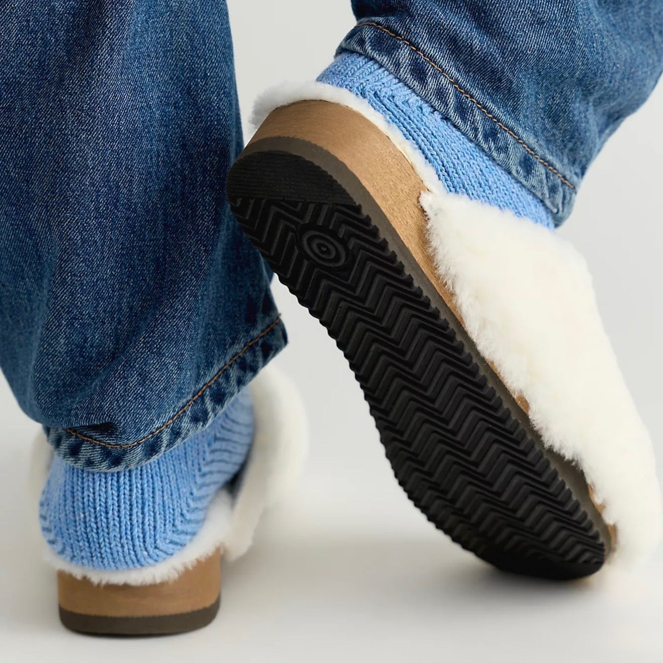 A close-up of a person wearing blue jeans and light blue knit socks with white faux-fur trim, standing on a light gray studio floor. One shoe is lifted to show the black treaded sole and tan footbed.