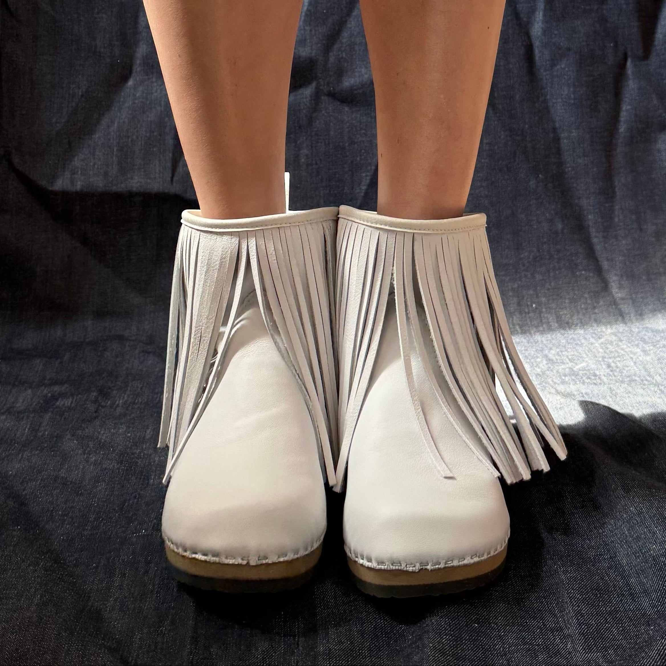 White fringe ankle boots worn by a model, shown from the front against a dark blue fabric backdrop.