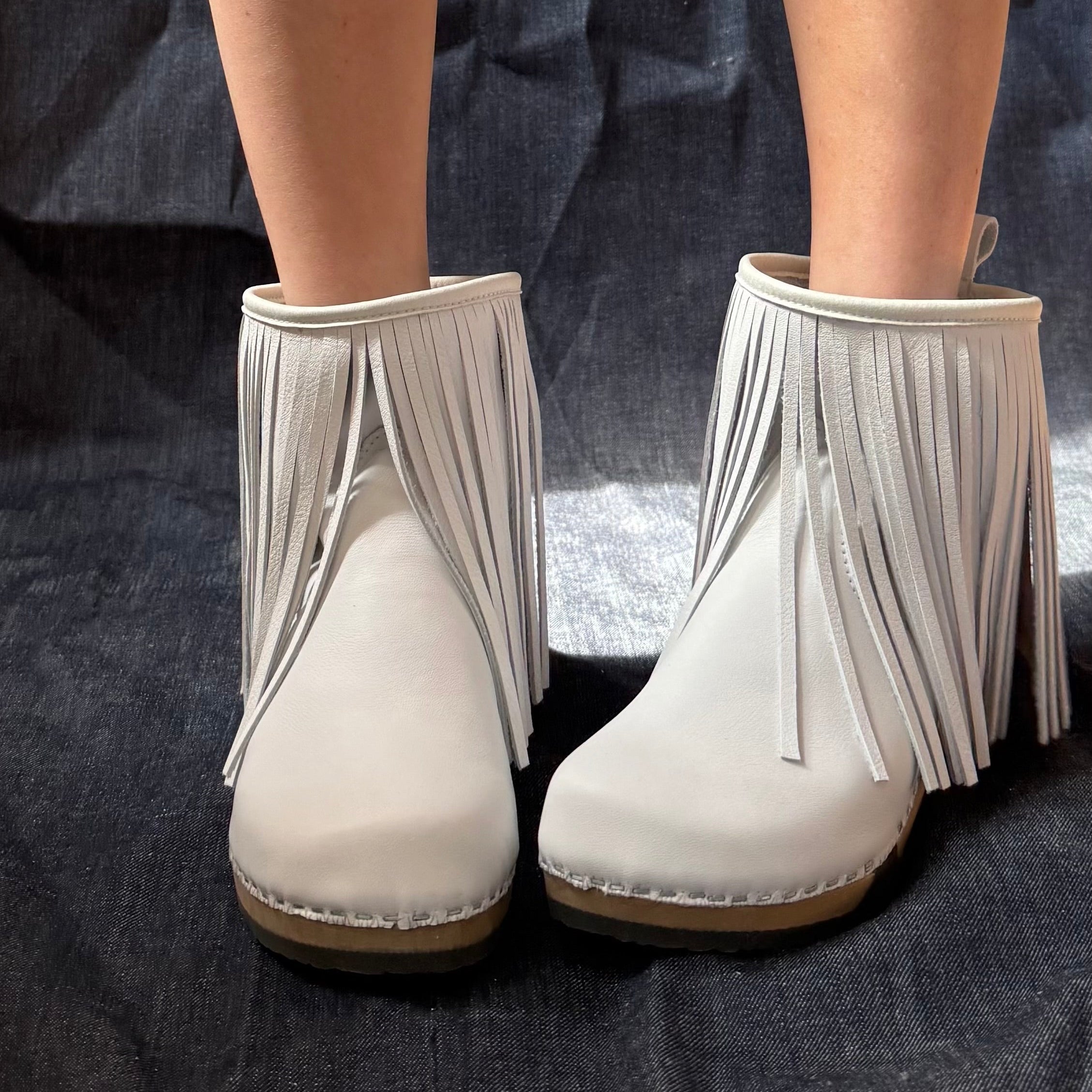 A pair of off-white leather ankle boots with long fringe detailing, shown on a model's feet against a dark textured fabric background.