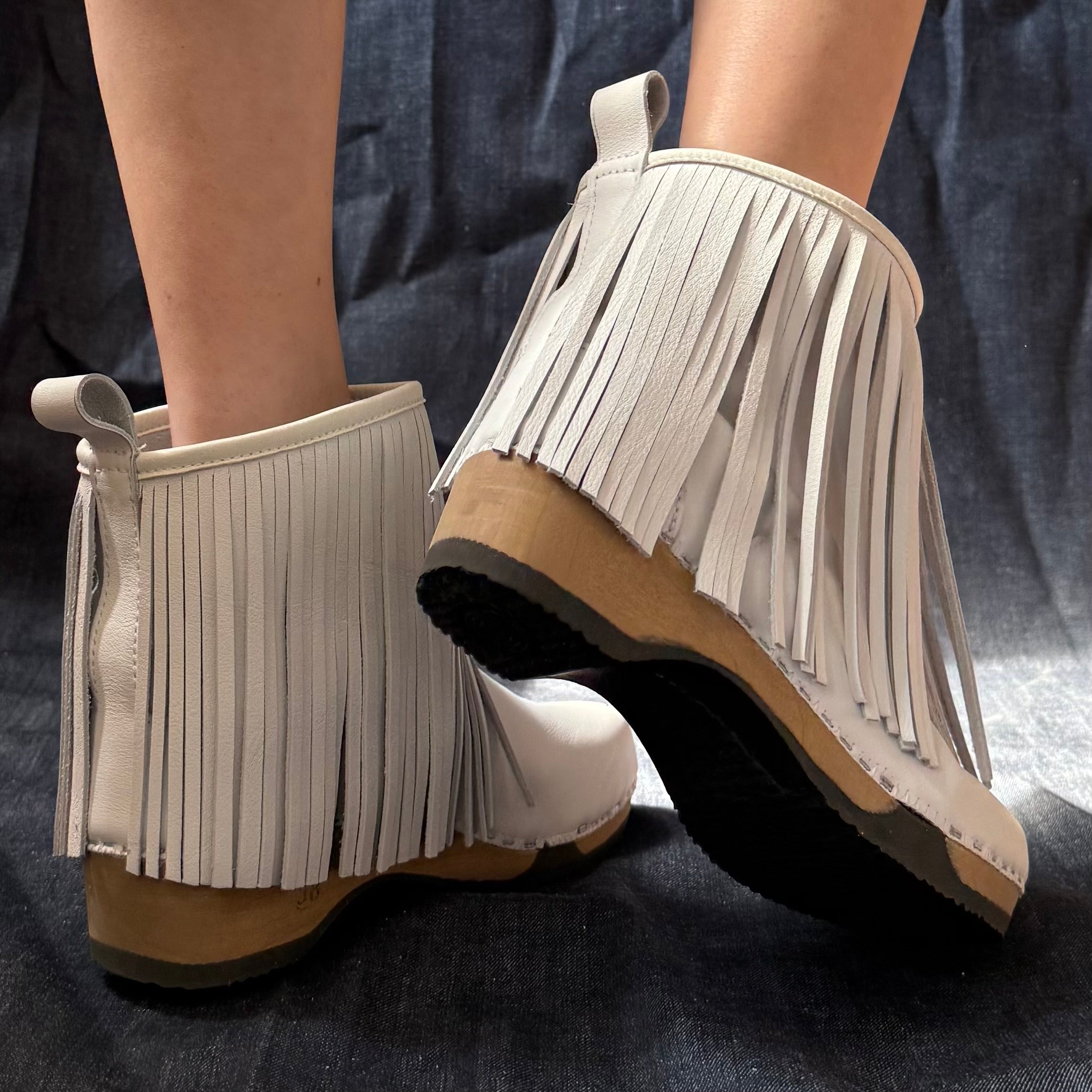 A pair of off-white fringed ankle boots with tan and black wedge soles, shown being worn on a person's feet against a dark blue fabric background.
