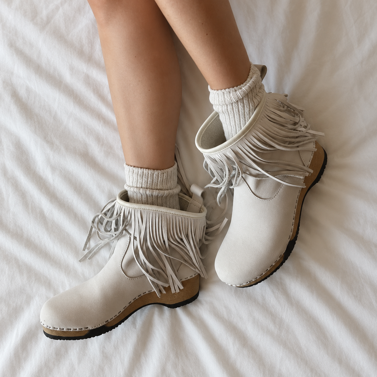 A pair of off-white fringed ankle boots with a wedge heel, shown on a model's feet with ribbed cream socks on a white bedsheet background.