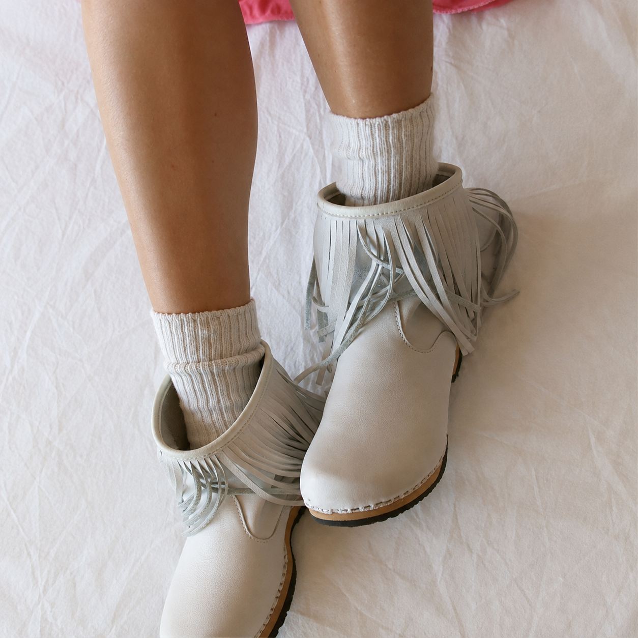 A person wearing light beige fringed ankle boots with white ribbed socks, standing on a white fabric surface.